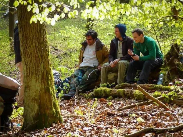 Drei Personen sitzen im Wald auf dem Boden und halten Stöcke in der Hand