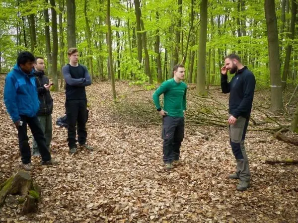 Fünf Personen stehen im Wald und diskutieren in einer Laublandschaft