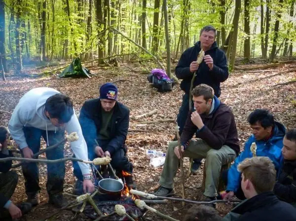 Gruppe bereitet Essen über Feuer in einem Waldgebiet mit Kochtopf und Stockbrot
