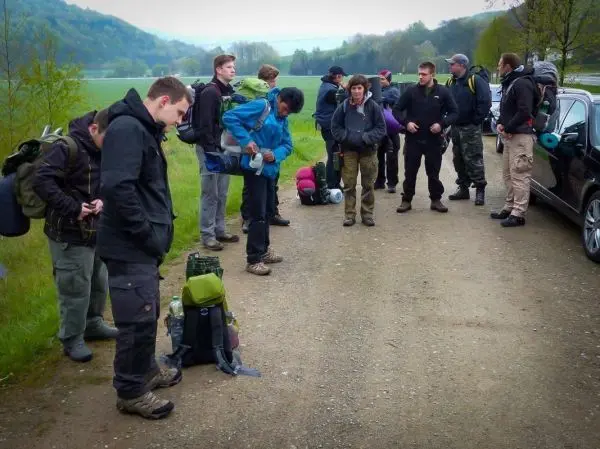 Gruppe mit Rucksäcken steht an einem Weg, einige überprüfen ihre Ausrüstung