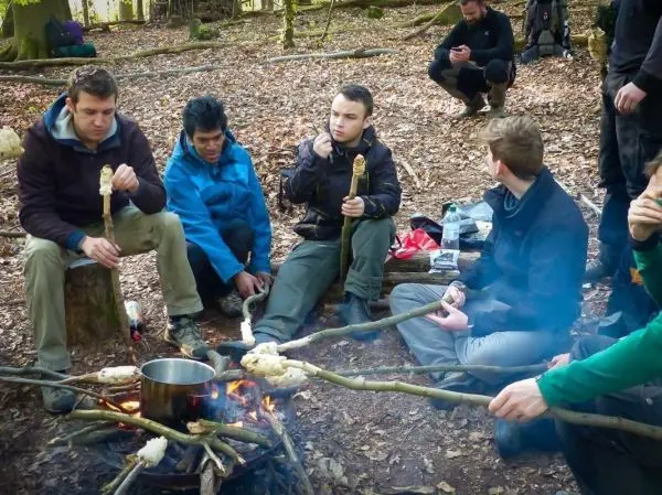 Gruppe sitzt um ein Feuer, während sie Essen auf Stockspießen zubereitet