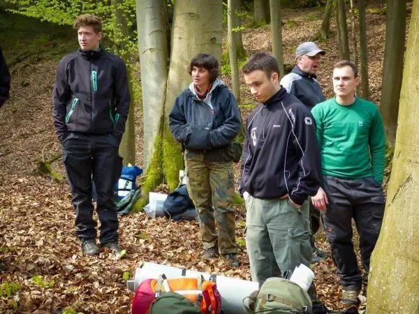 Gruppe steht im Wald auf Laub, Rucksäcke und Ausrüstung liegen am Boden
