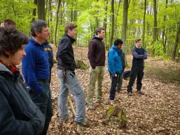 Gruppe von Personen steht im Wald auf Laubboden und hört aufmerksam zu