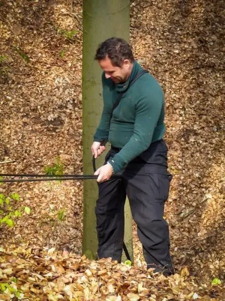 Mann hält zwei Stöcke in der Hand und steht auf Laubboden