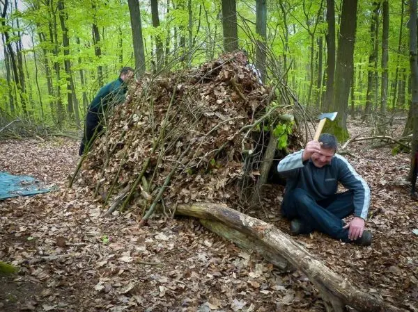 Mann mit Axt sitzt neben einer selbstgebauten Laubhütte im Wald
