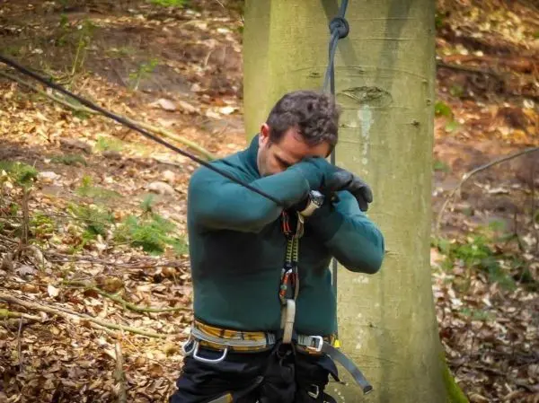 Mann mit Kletterausrüstung hängt an einem Seil, Arme vor dem Gesicht verschränkt