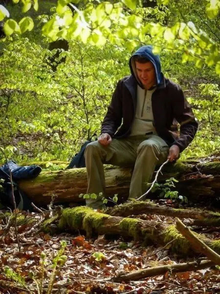 Mann sitzt auf einem umgestürzten Baumstamm und bearbeitet einen Stock