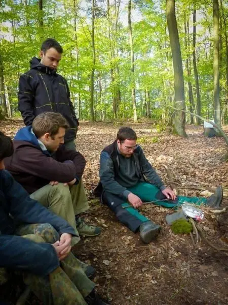 Mann sitzt vor einem kleinen Bodenfeuer und bereitet Zunder vor, während andere zuschauen