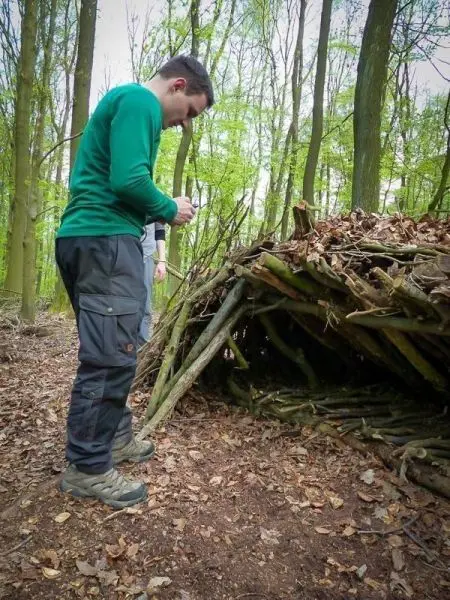 Mann steht neben einer selbstgebauten Laubhütte im Wald und hält ein Werkzeug