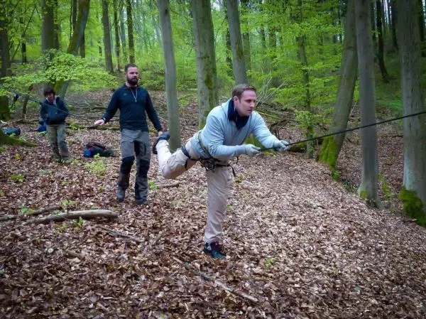 Mann überquert selbstgebaute Seilbrücke im Wald, während andere sichern