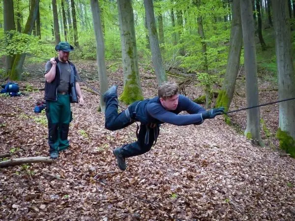 Mann überquert selbstgebaute Seilbrücke im Wald, während ein anderer zuschaut