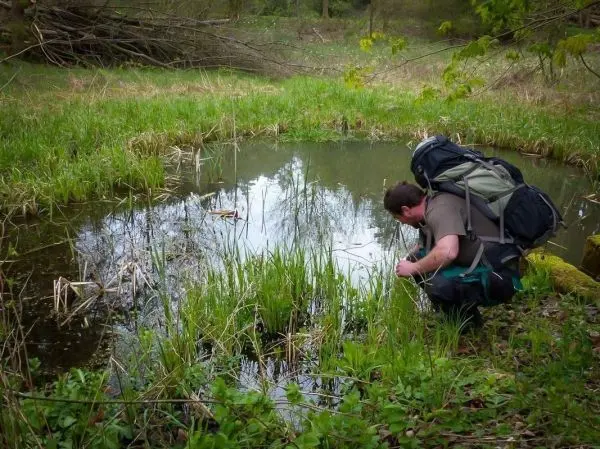 Person beugt sich über einen kleinen Teich mit Rucksack