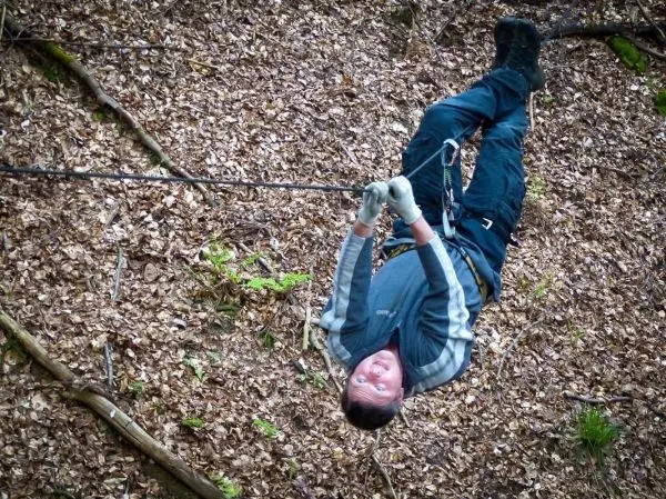 Person hängt kopfüber an einem Seil über Laubboden beim Überlebenstraining