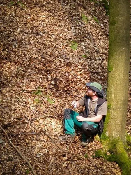 Person sitzt an Baumstamm und betrachtet eine Flasche im Wald
