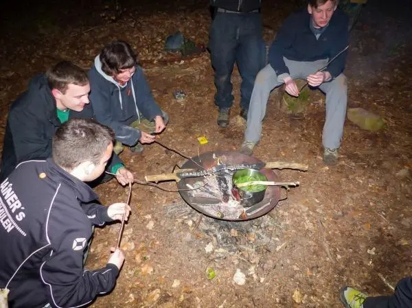 Personen halten Stöcke über eine Feuerstelle im Wald beim Überlebenstraining