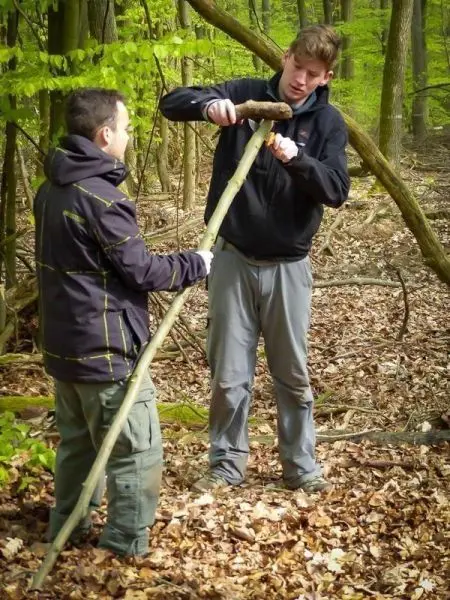 Zwei Personen bearbeiten einen langen Stock im Wald mit einem kürzeren Holzstück