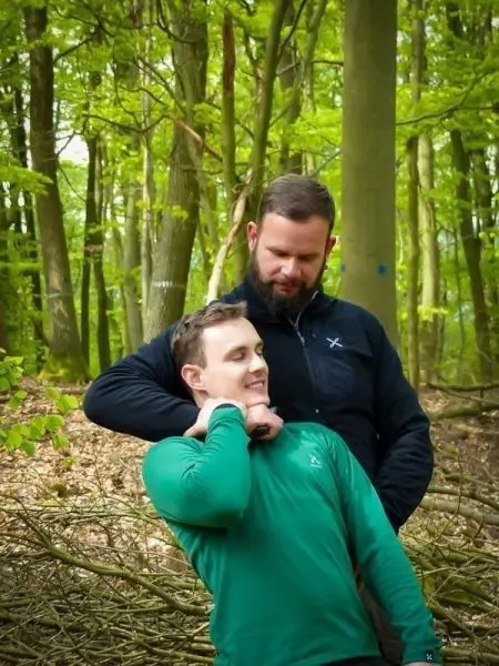Zwei Personen stehen in einem Wald, eine Person hält die andere am Hals