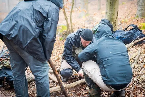 Drei Personen arbeiten an einer Holzstruktur im Wald, einer schnitzt mit einem Messer