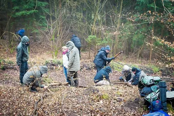 Gruppe arbeitet an Holzstücken im Wald, einige halten Stöcke, andere knien am Boden