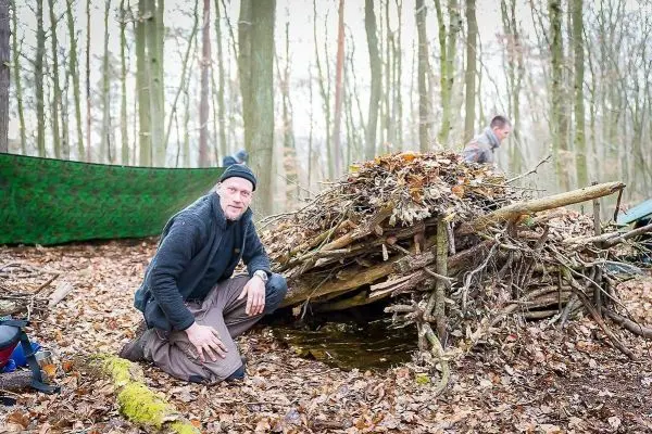 Mann kniet neben einer selbstgebauten Laubhütte im Wald
