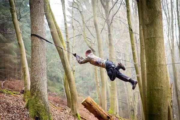 Mann überquert selbstgebaute Seilbrücke zwischen zwei Bäumen im Wald
