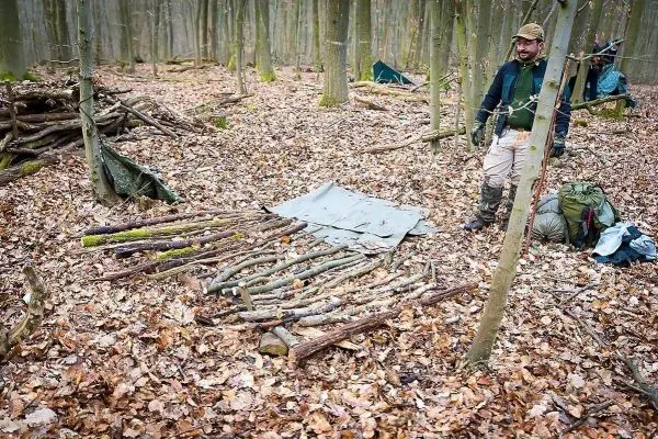 Person steht neben einer selbstgebauten Unterlage aus Ästen im Wald