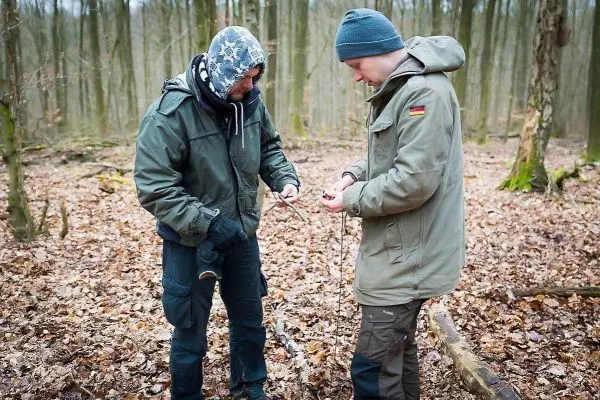 Zwei Personen arbeiten im Wald an einem Holzstück und halten Werkzeuge in der Hand
