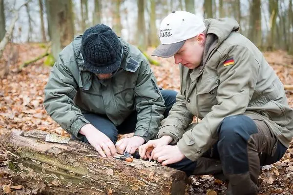 Zwei Personen bearbeiten einen Baumstamm mit einem Messer im Wald