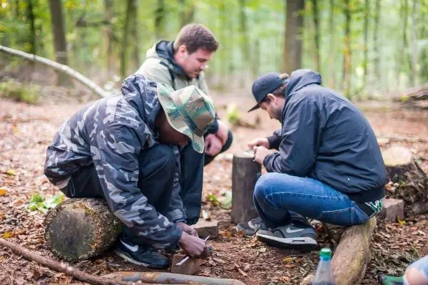 Drei Personen sitzen auf Baumstämmen und bearbeiten Holzstücke beim Überlebenstraining