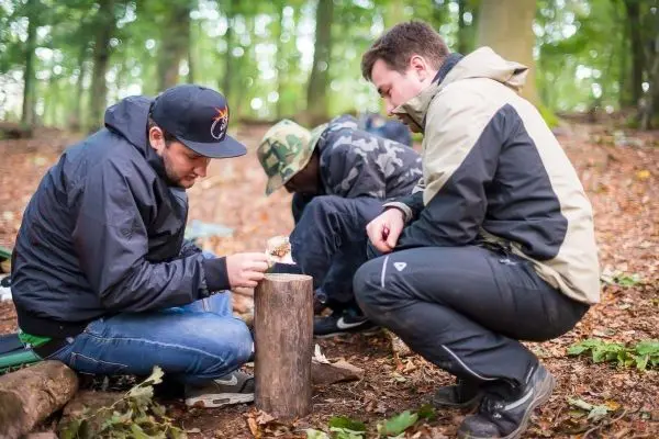 Drei Personen sitzen im Wald und bearbeiten einen Holzstock beim Überlebenstraining