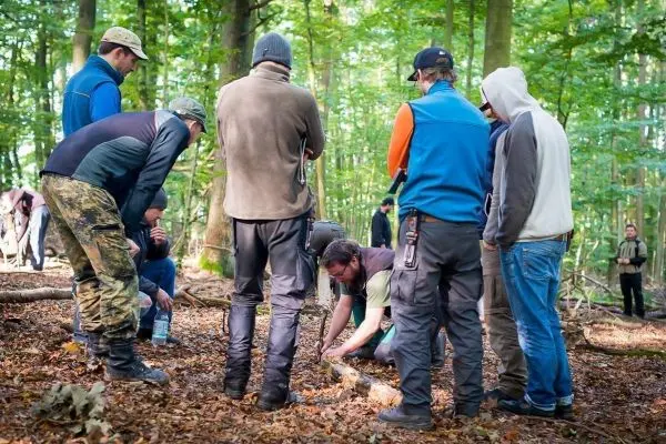 Gruppe beobachtet Holzarbeiten im Wald, einer bearbeitet einen Stock am Boden