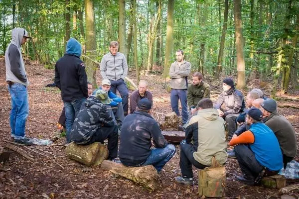 Gruppe sitzt um ein kleines Feuer auf dem Waldboden, Holzscheite als Sitzgelegenheit