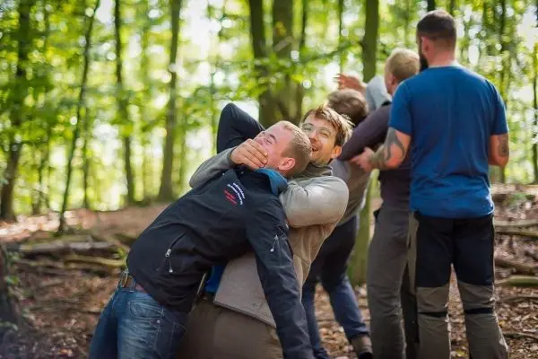 Gruppendynamik im Wald, eine Person hält einen anderen fest, während weitere Teilnehmer im Hintergrund stehen