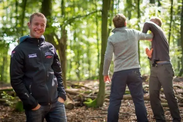 Lächelnder Mann steht im Wald, während zwei Personen eine Auseinandersetzung simulieren