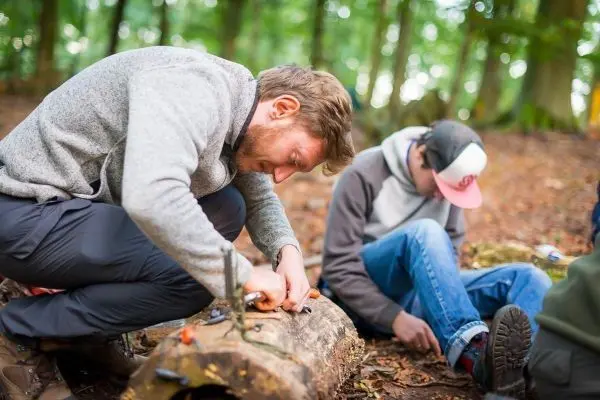 Messer bearbeitet Holzstück im Wald, während eine Person sitzt und zuschaut