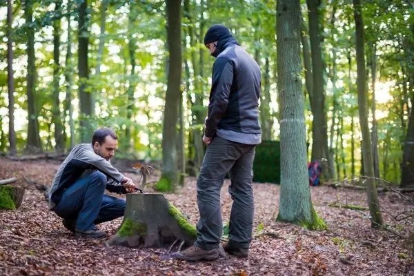 Person sitzt auf Baumstumpf und zündet Feuer an, Person steht daneben