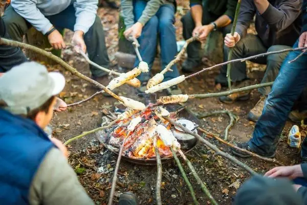 Stöcke mit Teig über offenem Feuer geröstet, umgeben von Personen im Wald