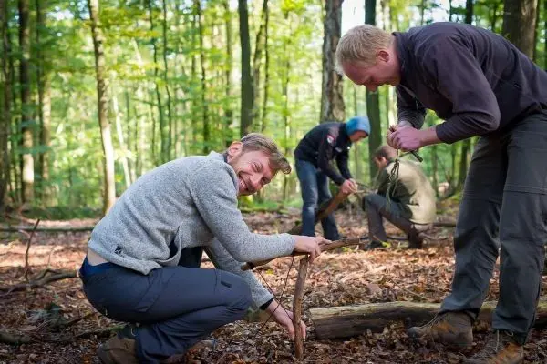 Zwei Personen arbeiten an einer Holzstruktur im Wald, während andere in der Nähe beschäftigt sind