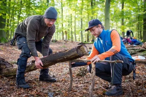 Zwei Personen bearbeiten einen Baumstamm im Wald beim Überlebenstraining
