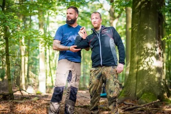 Zwei Personen demonstrieren eine Technik im Wald, eine Person hält die andere am Arm