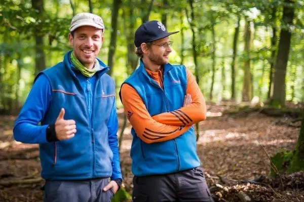 Zwei Personen in blauer und orangefarbener Jacke stehen im Wald