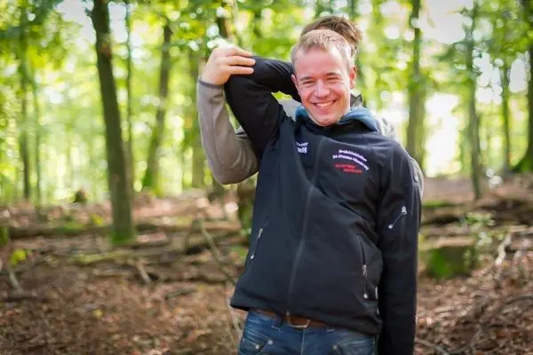 Zwei Personen stehen im Wald, eine Person umarmt die andere von hinten