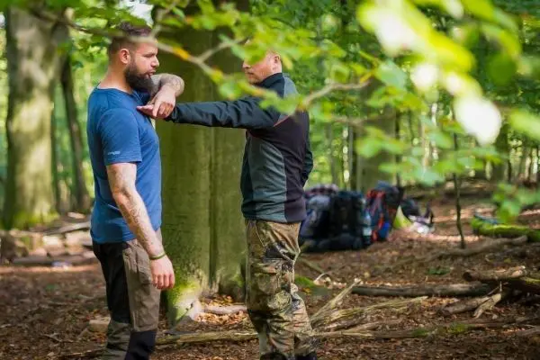 Zwei Personen üben eine Technik im Wald, während Rucksäcke im Hintergrund stehen
