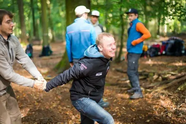 Zwei Personen ziehen an einem Seil im Wald, während andere im Hintergrund stehen