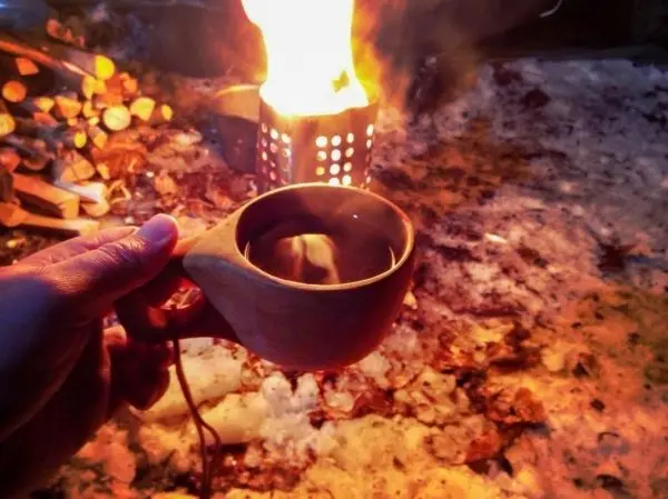 Hand hält eine Tasse vor einem Feuer im Freien