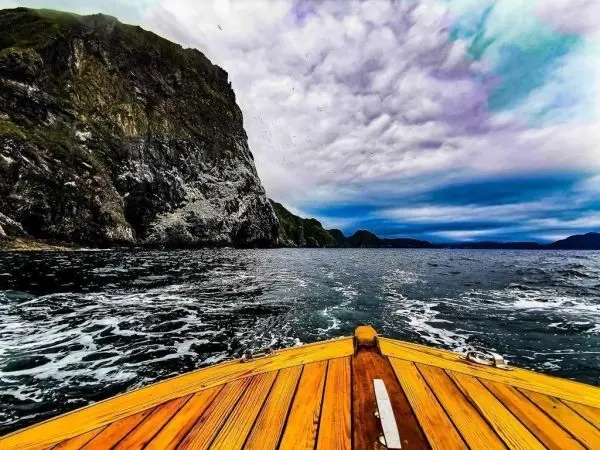 Holzboot fährt auf unruhiger See in Richtung steiler Klippen unter bewölktem Himmel