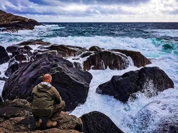 Person sitzt auf Felsen am Meer, Wellen schlagen gegen die Steine