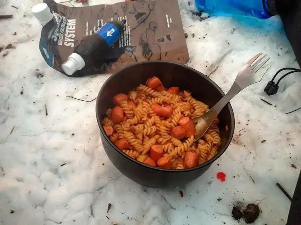 Schüssel mit Nudeln und Würstchen auf Schnee, daneben Wasserflasche und Verpackung