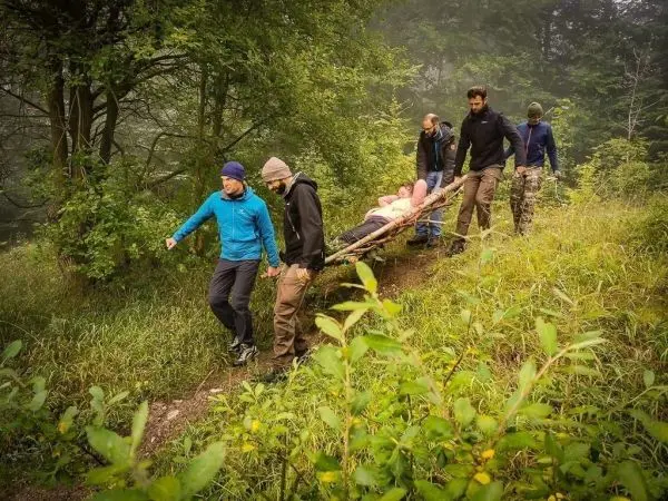 Sechs Personen tragen eine Trage mit einer liegenden Person durch bewaldetes Gelände
