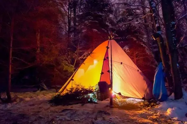 Tarp-Notunterkunft leuchtet orange im Dunkeln, umgeben von Schnee und Bäumen